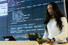 A woman with a laptop stands in front of a large screen displaying computer code.